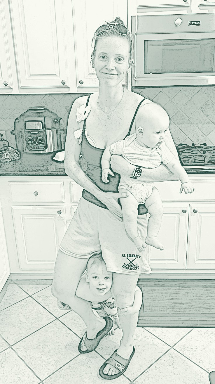 Kate in her kitchen, holding her youngest with a toddler wrapped around her leg.
